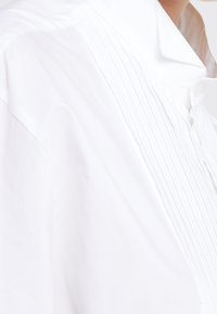 Vit skjorta med veckad design på framsidan, knappstängning och en strukturerad krage. Tyget verkar vara mjukt och lätt.