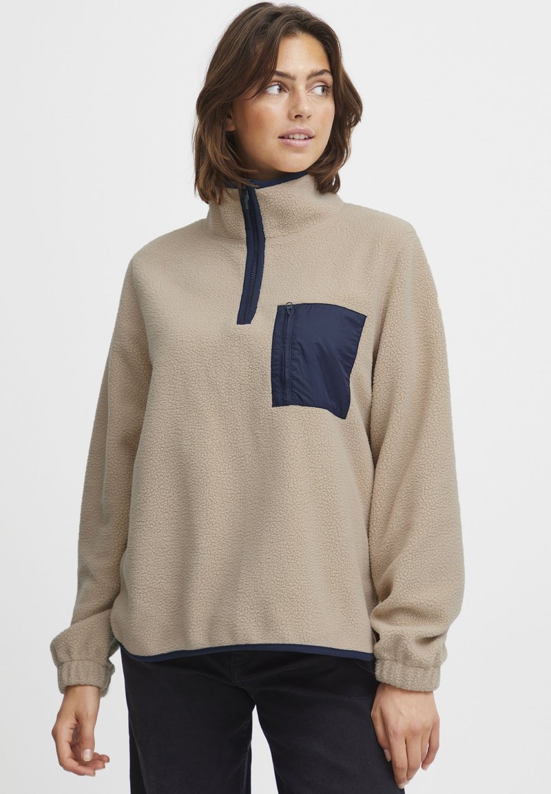 Beige fliisist pullover, millel on tumesinine lukugaTasku. Omab kõrget kaelust ja mansetiga varrukasid, pakkudes tekstuurset ja pehmet välimust.