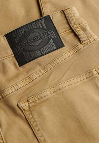 Tela de algodón beige con costuras visibles, que presenta una etiqueta de cuero negro en relieve con la inscripción "SUPERDRY STANDARD DENIM GOODS."