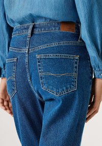 Modré džínové kalhoty s dvěma zadními kapsami, kontrastním prošíváním a hnědou koženou etiketou na pase. Měkká textura a uvolněný střih.