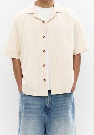 Mand iført en cremefarvet kortærmet knappet skjorte over en hvid t-shirt med lyseblå denimbukser, hænderne i lommerne.
