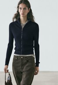 Navy ribbed zip-up cardigan with a fitted silhouette, worn over a grey shirt. Paired with dark brown pants and a small handbag.