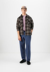 Jonge man staand, draagt een beige pet, een bruin geruite jas over een lichtpaarse shirt, blauwe broek en zwarte sneakers tegen een witte achtergrond.