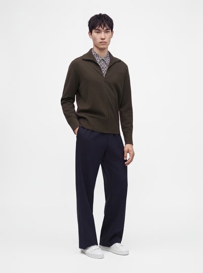 Jeune homme portant un pull zippé vert foncé par-dessus une chemise à motifs, un pantalon large bleu marine et des baskets blanches, debout devant un fond uni.