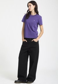T-shirt di cotone viola con scollo rotondo. Abbinato a jeans neri a gamba larga e vita alta. Modella in piedi con le mani in tasca su uno sfondo chiaro.