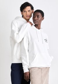 Sudaderas blancas de tela suave, con bolsillos frontales y cordones. Una tiene un pequeño detalle gráfico en negro. Los modelos están de pie muy cerca uno del otro.