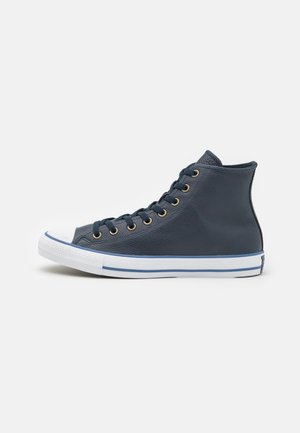 Zapatilla deportiva de caña alta en azul marino con suela de goma blanca, ralla azul y cordones negros sobre un fondo liso y claro.