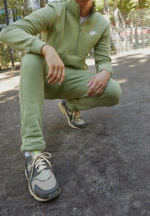 Personne portant un survêtement vert Nike et des baskets grises accroupie sur un chemin de terre avec des arbres et une clôture en arrière-plan.