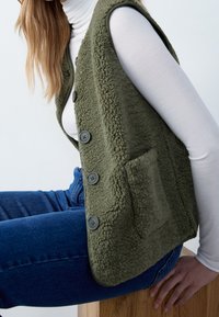 Person, der auf einem Holzblock sitzt, trägt ein weißes Langarmshirt, eine olivgrüne strukturierte Weste mit Knöpfen und blaue Jeans vor einem schlichten Hintergrund.