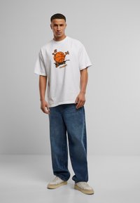 Hvit oversized t-skjorte med grafikk av en basketball og en tegneseriefigur. Kombinert med vide blå jeans og beige joggesko.
