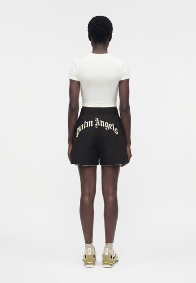 Άτομο που στέκεται με την πλάτη προς τα έξω, φοράει εφαρμοστή λευκή crop top, μαύρα σορτς με το κείμενο «Palm Angels» και μπεζ αθλητικά με λευκές κάλτσες.