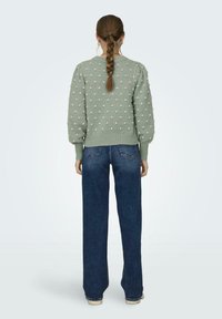 Groen gebreid sweater met getextureerde roze stippen, met geribde manchetten en zoom. Gecombineerd met brede blauwe denim jeans, gezien van achter.