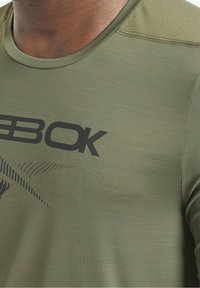 Olivově zelené sportovní tričko s kulatým výstřihem a černým logem. Má jemnou texturu a nenápadný vzor na látce.