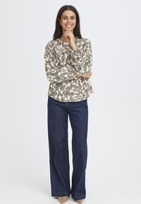 Femme souriante se tient avec une main près du menton, portant un blouse à manches longues à motifs, un jean large noir et des chaussures beige à bouts pointus sur fond blanc.