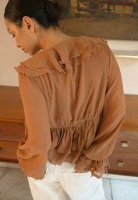 Bruine, gerimpelde blouse met lange mouwen en een geplooide taille. Textuurstof met delicate kantaccenten op de mouwen. Draag het met witte broeken.