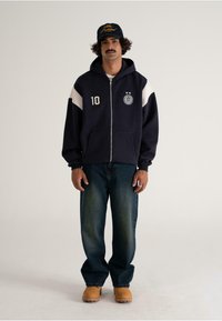 Man with dark curly hair and mustache wears navy hoodie with white patches, blue jeans, tan boots, and black cap, standing against plain background.