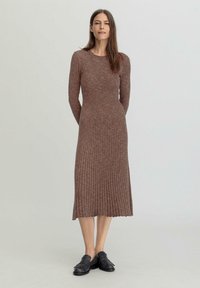 Langes, geripptes Strickkleid in brauner Farbe mit Rundhalsausschnitt, taillierter Passform und schwingendem Rock. Kombiniert mit schwarzen Slippern.