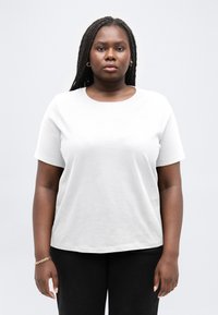 Νεαρή γυναίκα με μακριά κοτσίδα, φορώντας λευκό κοντομάνικο πουκάμισο, μαύρο παντελόνι, χρυσούς κρίκους στα αυτιά και βραχιόλι, στέκεται μπροστά σε ανοιχτό φόντο.