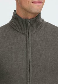 Mann trägt einen grauen, gerippten Strickpullover mit hohem Kragen und einem teilweise zugeknöpften Reißverschluss vor einem hellen Hintergrund.