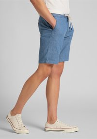 Shorts de chambray azul con cinturilla elástica y cordón, que cuentan con bolsillos laterales y un ajuste relajado. Llevados con zapatillas beige.