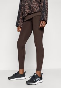 Hnedé legíny s priliehavým dizajnom, so švami na bokoch a hladkou textúrou. Kombinované s čiernymi športovými topánkami a topom s leopardím vzorom.