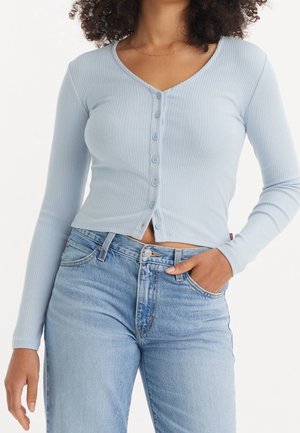 Femme portant un cardigan côtelé bleu clair boutonné et un jean bleu clair taille haute, avec une main dans la poche.