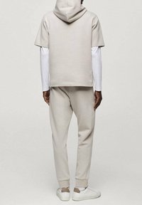 Man wearing beige short-sleeve hoodie over a white long-sleeve shirt with matching beige joggers and white sneakers, standing facing away.