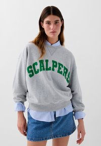 Grå sweatshirt med "SCALPERS" i grøn skrift, lagt over en blåstribet skjorte, parret med en denim miniskørt.