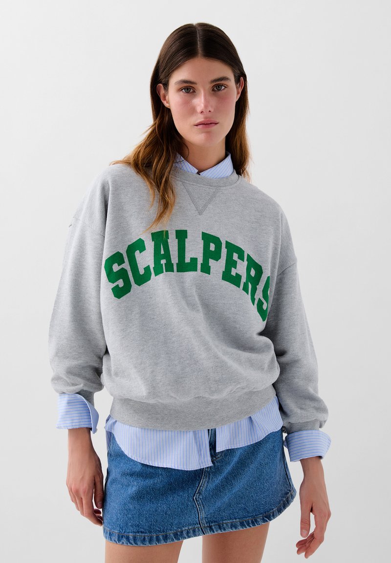Grå sweatshirt med "SCALPERS" i grøn skrift, lagt over en blåstribet skjorte, parret med en denim miniskørt.