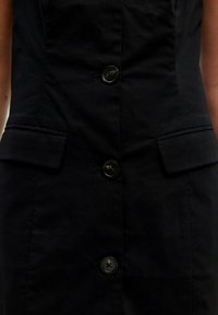 Schwarzes Kleid mit figurnaher Passform, sechs Knöpfen an der Vorderseite und zwei aufgesetzten Taschen an den Seiten. Der Stoff wirkt glatt und leicht strukturiert.