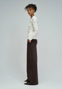 Donna in piedi di lato, che indossa una maglietta bianca a maniche lunghe, pantaloni larghi scuri e scarpe con la punta a vista, su uno sfondo chiaro uniforme.