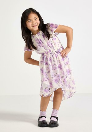 Ung pige i en lilla blomsterkjole, lyserøde sokker og sorte sko står med hænderne på hoften, smilende og læner sig let fremad.