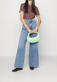 Groene, gebogen tas met een zachte textuur, gecombineerd met een rode shirt met zigzagpatroon en lichtblauwe wijde jeans. Zwarte hakken.