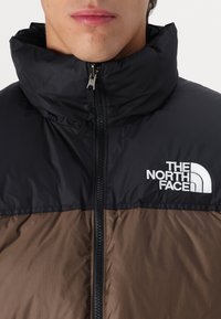 Jachetă puf neagră și maro cu guler înalt, fermoar frontal, logo The North Face pe pieptul stâng, material rezistent la apă și textură matlasată.