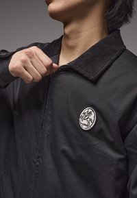 Μαύρο σακάκι με γιακά από κοτλέ, με κυκλικό κεντητό σχέδιο με άλογο και τη λέξη "FORMER." Ελαφρώς ανοιχτό με φερμουάρ.