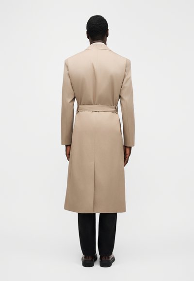 Beiger Trenchcoat mit Gürtel, langen Ärmeln, taillierter Form und einem einzelnen Rückschlitz, kombiniert mit schwarzer Hose und Schuhen.