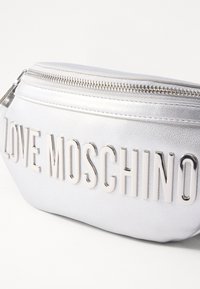Srebrna sintetična usnjena torbica z embossiranim napisom "LOVE MOSCHINO", zadrgo in gladko teksturo. Kompaktna ovalna oblika.