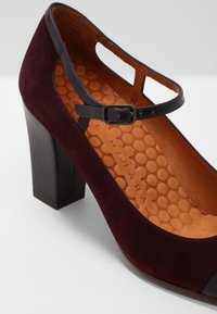 Chaussure Mary Jane en daim bordeaux foncé avec talon carré, bride noire avec boucle, et semelle intérieure texturée beige avec motif circulaire.