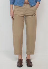 Chino beige con vestibilità rilassata, caratterizzati da una texture liscia, orli arrotolati e una cintura intrecciata marrone. Abbinati a mocassini marroni scuri.