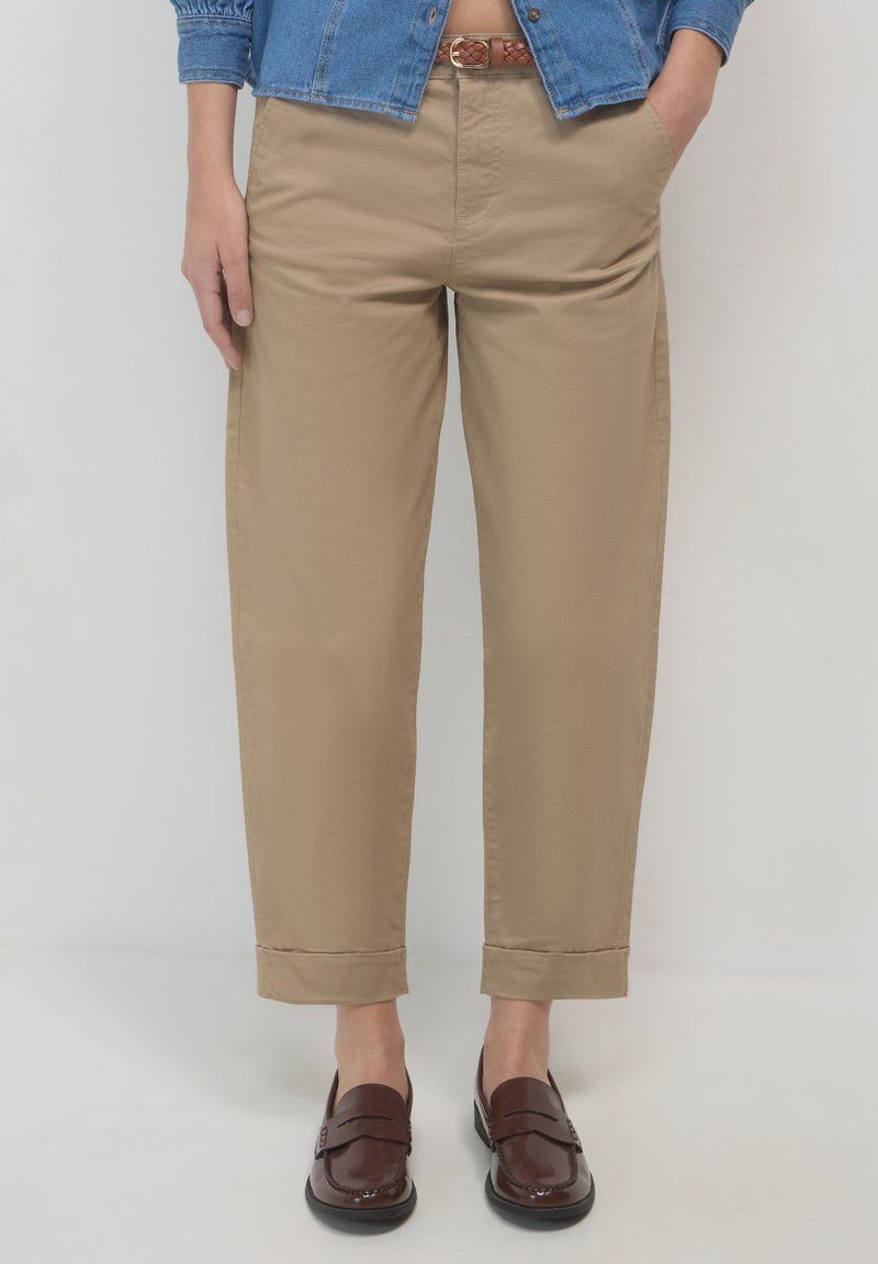 Chino beige con vestibilità rilassata, caratterizzati da una texture liscia, orli arrotolati e una cintura intrecciata marrone. Abbinati a mocassini marroni scuri.