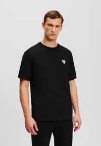 Tricou din bumbac negru cu guler rotund și mâneci scurte, având o mică grafică cu un inimioară albă pe piept.