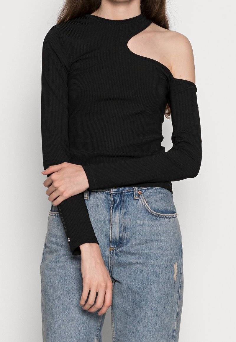 Schwarzer, ribbeliger Langarm-Top mit einem einzigartigen asymmetrischen Ausschnitt an der Schulter, kombiniert mit hellblauen Jeans.