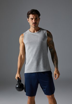 Grijze mouwloze sporttop met een ronde halslijn, gecombineerd met marineblauwe short. Het onderwerp houdt een zwarte kettlebell vast, waarbij gespierde armen en tattoos worden getoond.