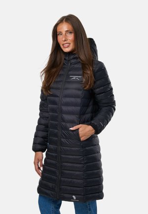 Femme portant un long manteau matelassé noir à capuche avec fermeture éclair devant et les mains dans les poches, debout devant un fond clair uni.