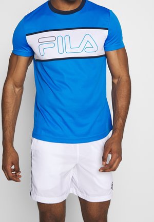 Mann trägt blaues FILA-T-Shirt mit weißem horizontalem Streifen und weiße Shorts, steht mit entspannten Armen seitlich am Körper.