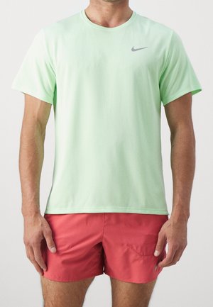Uomo che indossa una maglietta sportiva Nike a maniche corte verde menta e pantaloncini sportivi rossi, in piedi davanti a uno sfondo bianco uniforme.