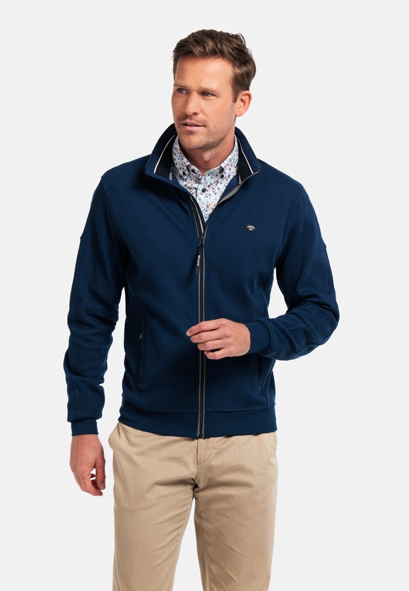 Navy zip-up sweatshirt made of soft fabric, featuring a high collar, side pockets, and contrasting inner trim. Worn over a patterned shirt.