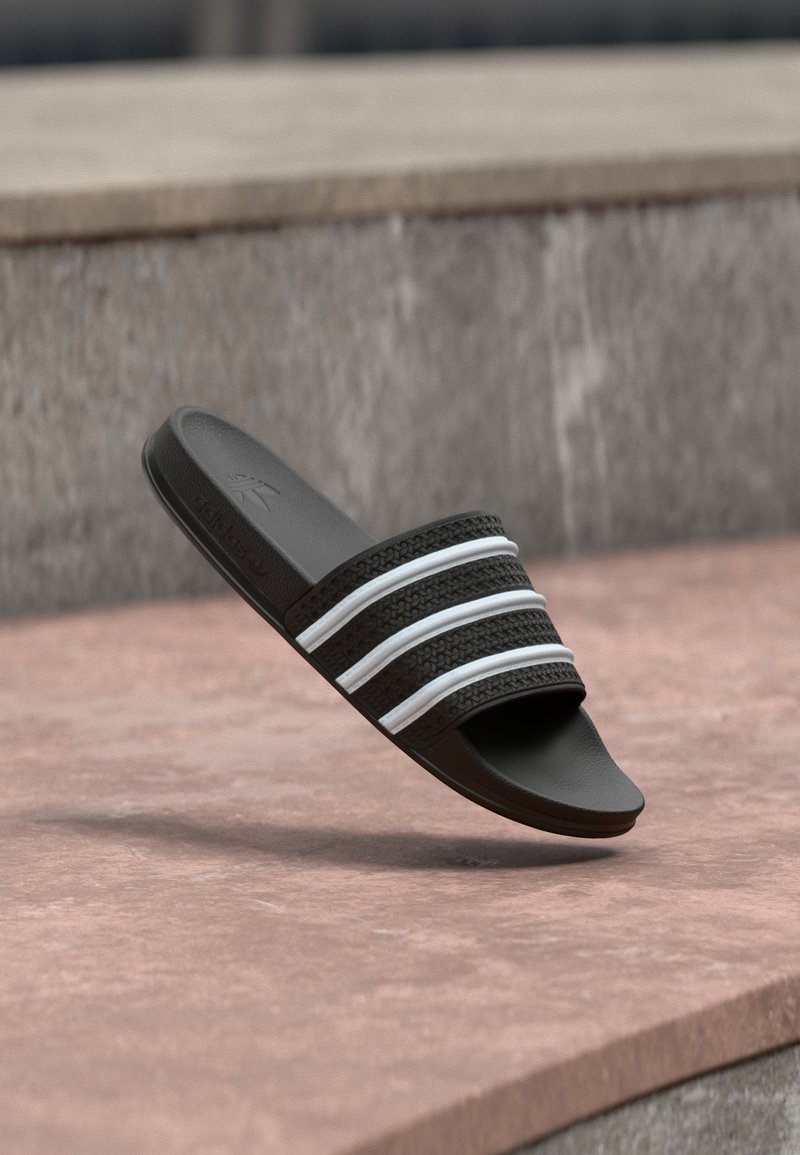 Sandale noire à slide avec des brides texturées ornées de trois bandes blanches, en matière caoutchoutée, avec une semelle plate et un design à bout ouvert.