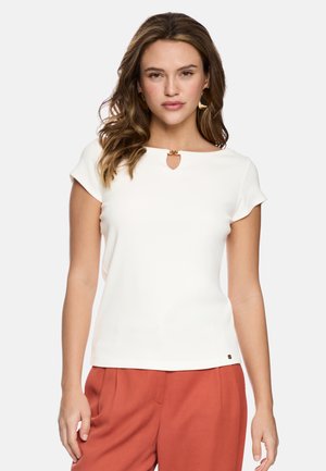 Woman with wavy brown hair wearing a white short-sleeve top with a small gold clasp at the neckline and rust-colored pants.