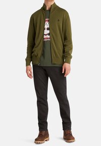 Sudadera de cremallera verde oliva con un cuello alto, llevada sobre una camiseta gráfica verde oscura, combinada con jeans negros y botas marrones.